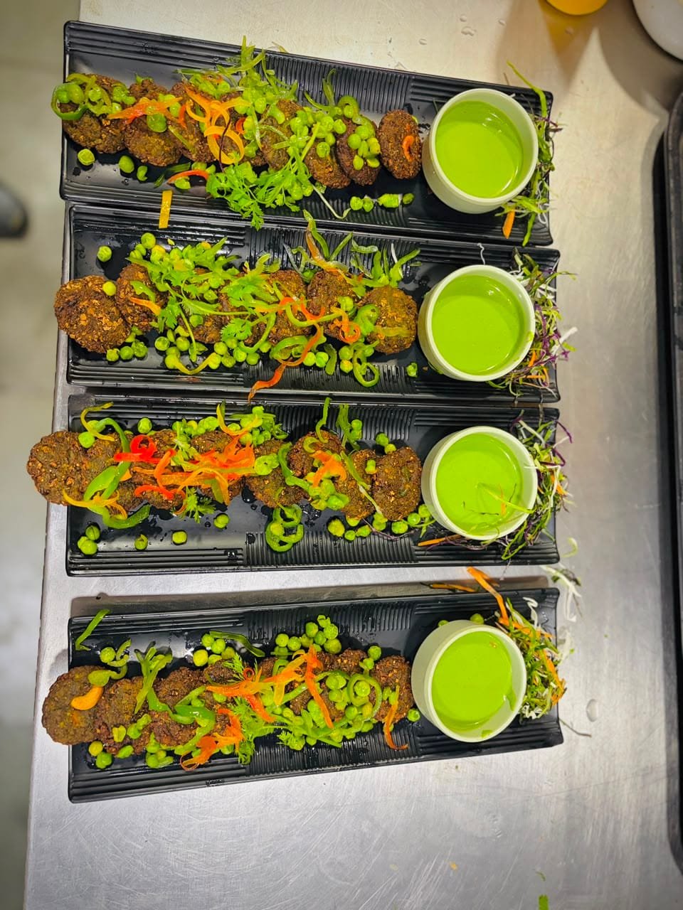 Four black platters of hara bhara kababs plated on the pass, each with green peas, ribboned pepper and a bowl of green chutney, ready for a banquet