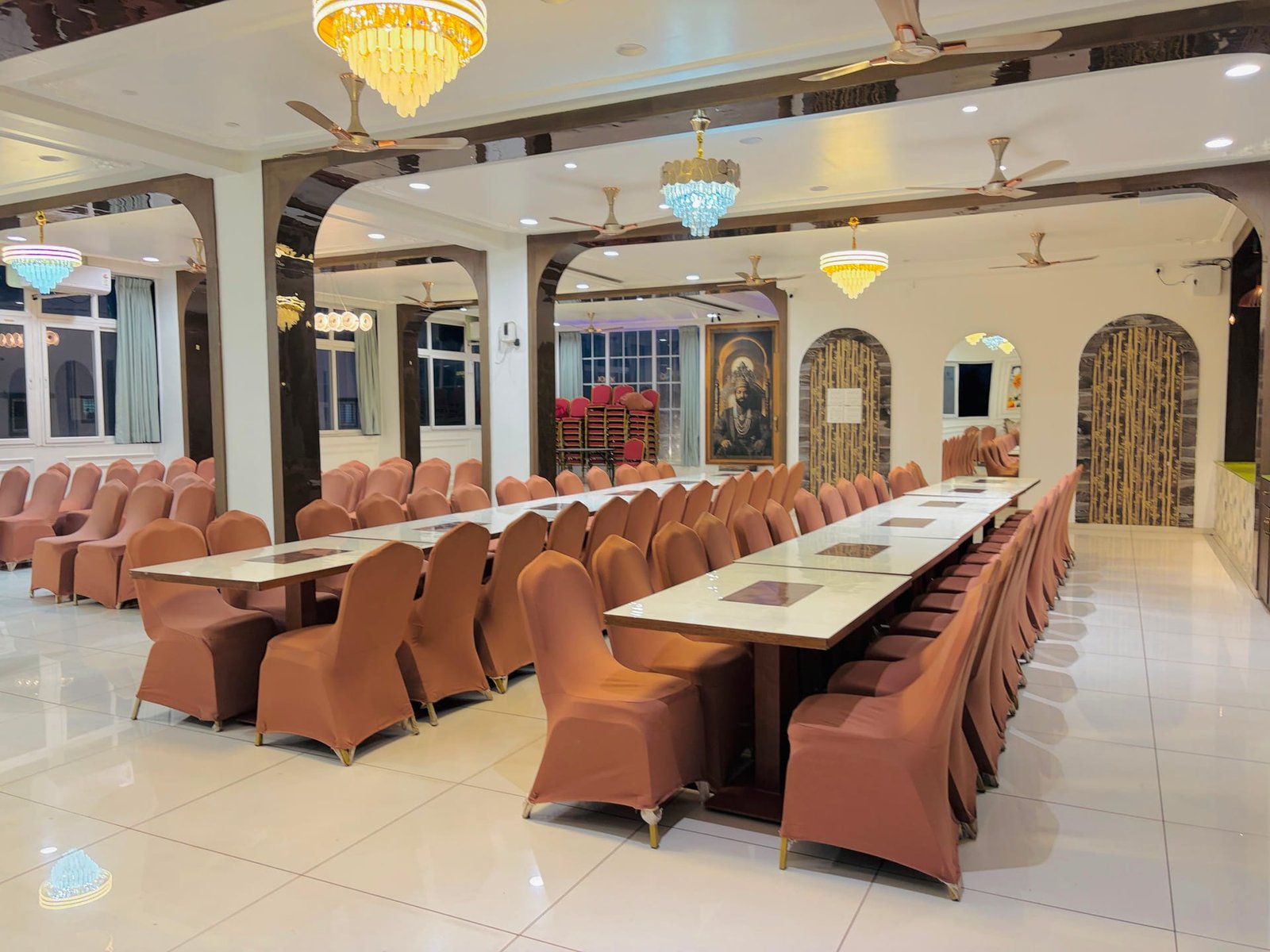 Long dining tables in the banquet hall dressed with marble-look tops and tan chair covers, arches leading through to the stage and a Shivaji Maharaj portrait on the far wall