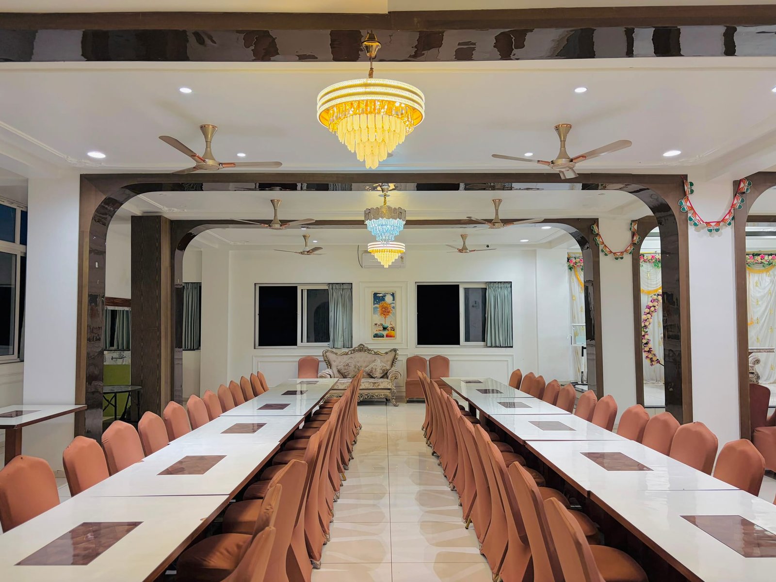 The dining hall shot down the length of two long tables dressed with pink chair covers and marble-look tops, framed by dark polished archways, with a golden crystal chandelier suspended overhead and a smaller blue chandelier behind, and a decorated stage visible at the far end