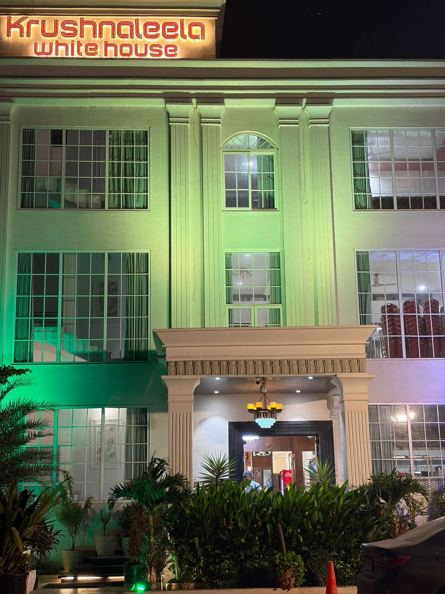 Close view of the columned main entrance beneath the neon Krushnaleela White House sign, with a chandelier glowing in the porch