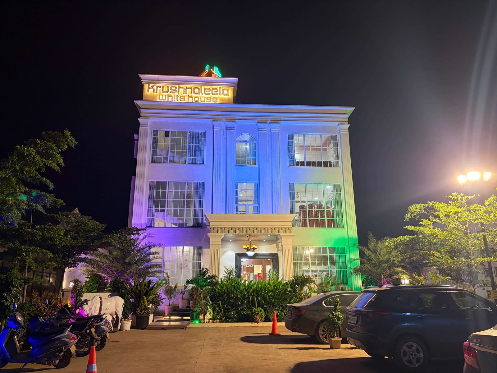 The three-storey white facade of Krushnaleela White House lit in blue and green at night, with the illuminated rooftop sign above the pillared entrance