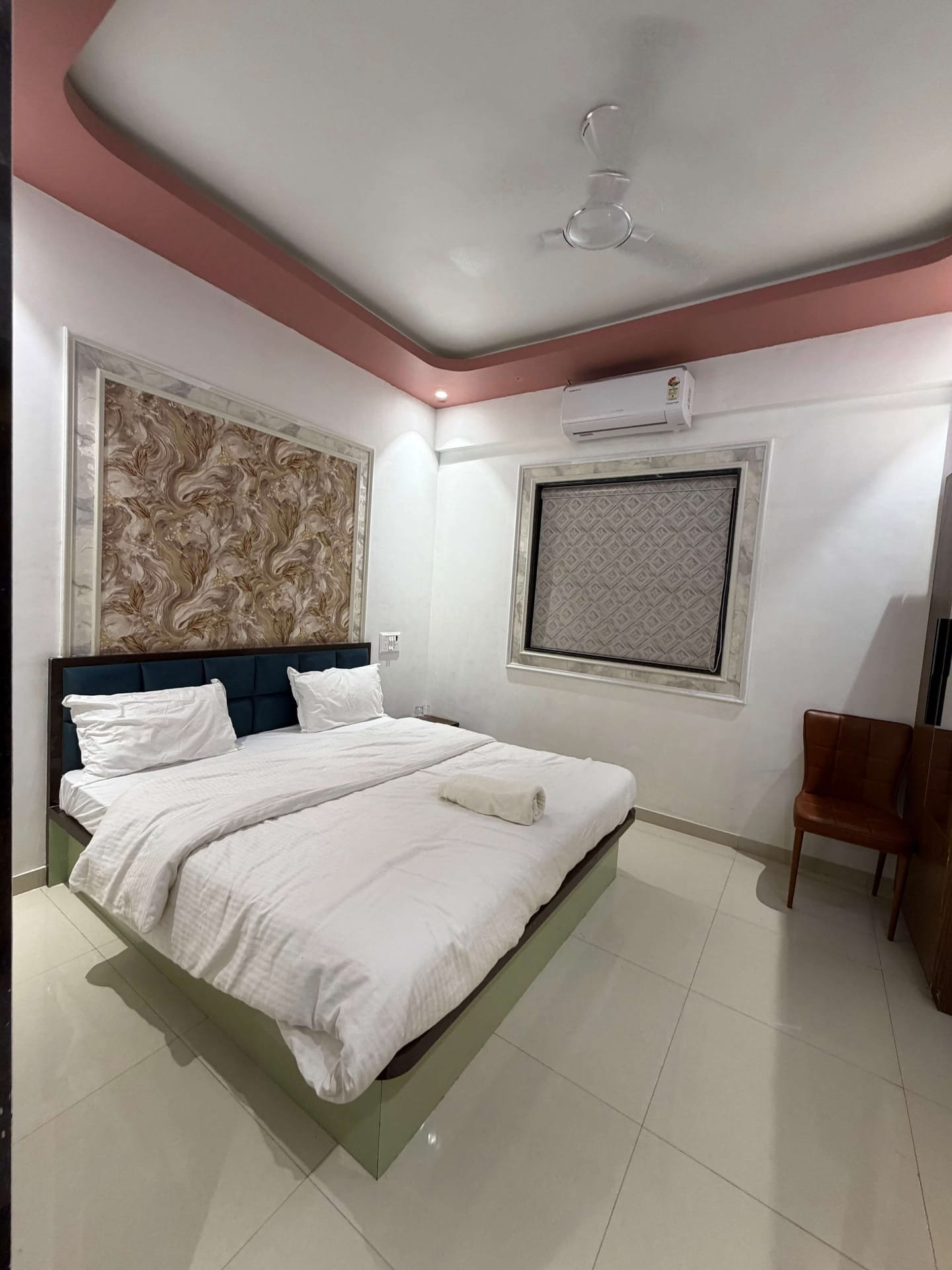 Deluxe AC room shot head-on — wide double bed in white linen on a sage-green bed base, tufted blue-velvet buttoned headboard against a marble-framed floral feature wall, split air conditioner above, chair and small side table alongside