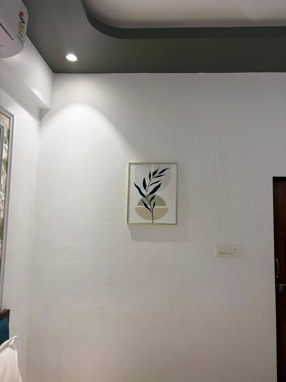 Framed botanical print on a plain white guest-room wall lit by a ceiling spotlight