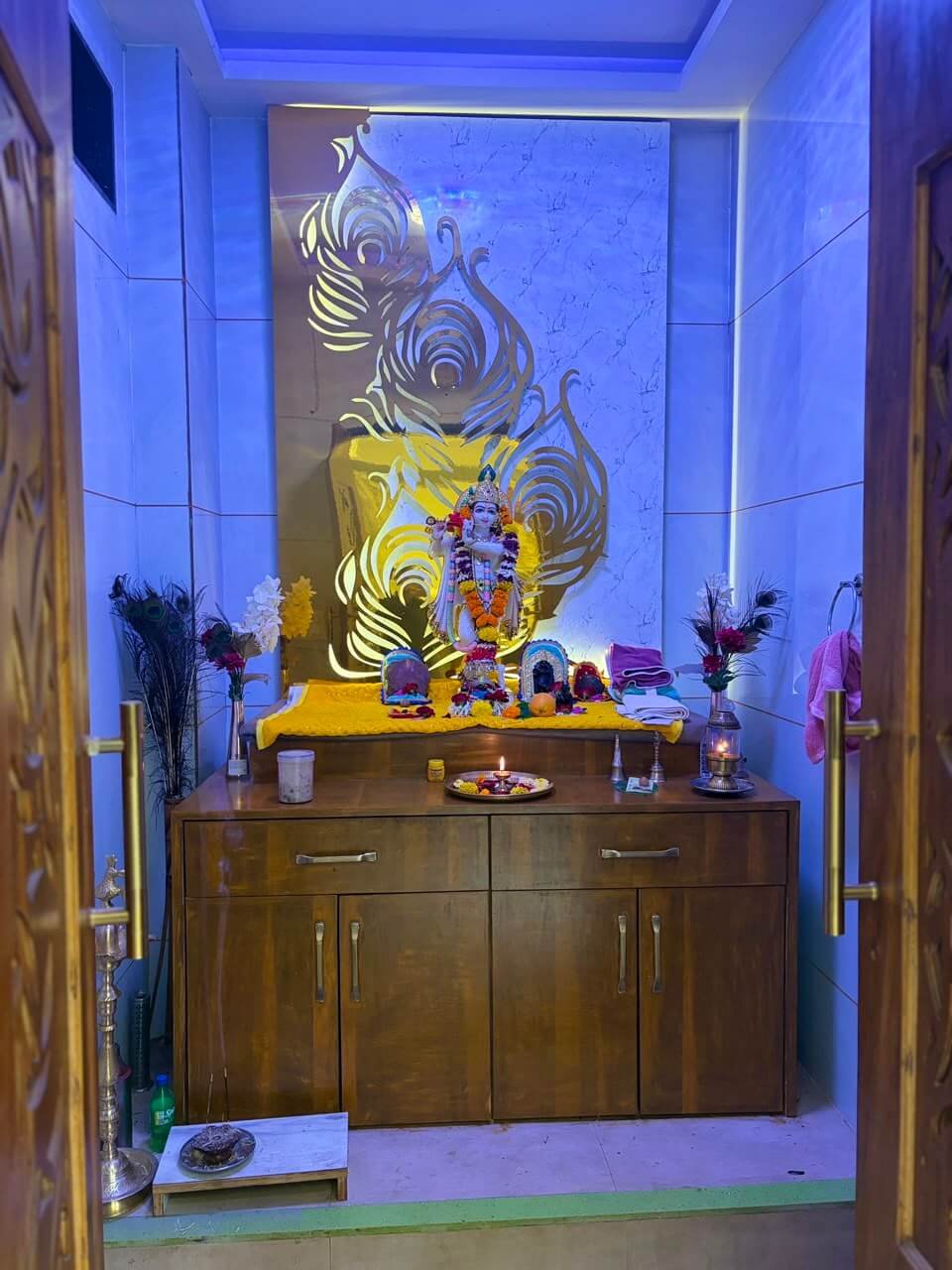 The house shrine — a garlanded Krishna murti before a gold peacock-feather cutout on marble, flanked by real morpankh, flowers and a lit diya, under soft blue cove lighting